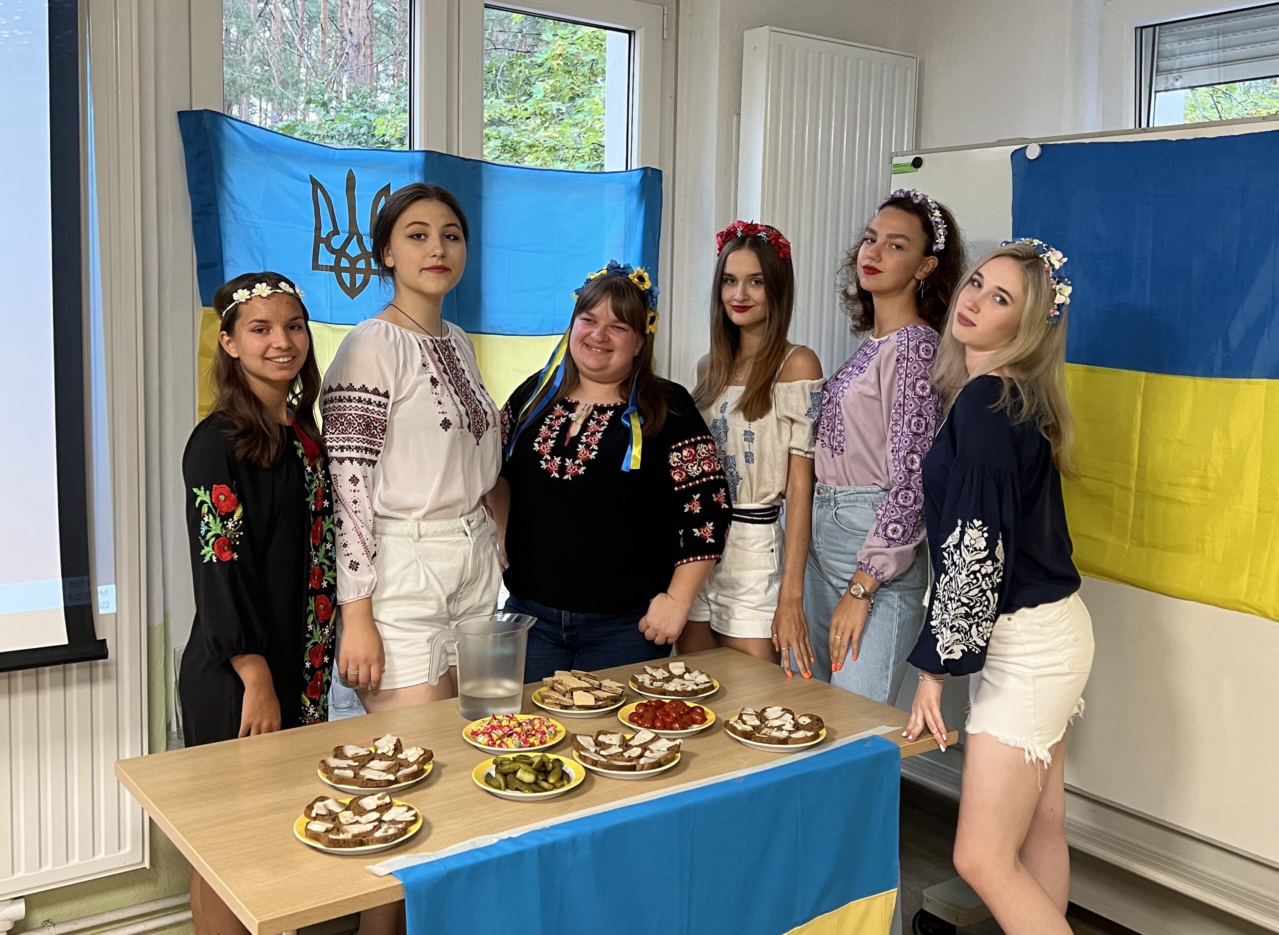 a group of young girls, some in Ukrainian folk cloths, behind a table with snacks, Ukrainian flags in the background