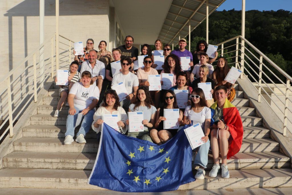 A group of young people sitting on stairs outside in bright sun, holding big EU flag